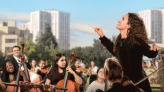 Bildbeschreibung: Filmplakat von Divertimento. Eine Frau dirigiert ein Orchester im Freien. Im Hintergrund sind hohe Gebäude und ein paar Bäume zu sehen.